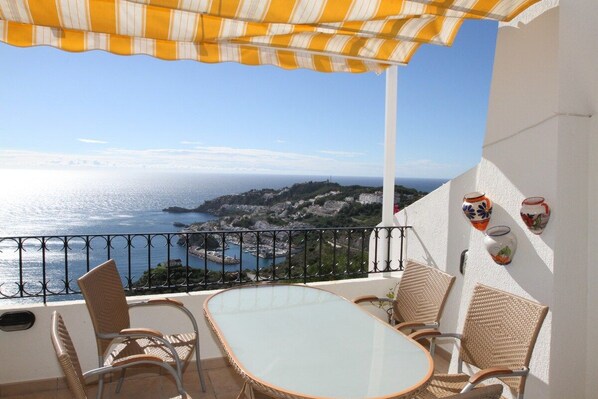 Un balcón con sillas de mimbre y una mesa, con vistas a la costa, edificios y el mar.