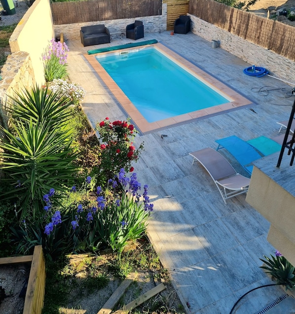 A rectangular swimming pool with a tiled deck, surrounded by a low brick wall. There are two lounge chairs and a small table with a chair. The area is landscaped with various plants and flowers.