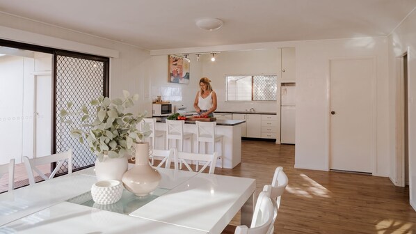 Uma cozinha moderna com armários brancos, uma área de jantar com mesa e cadeiras brancas, e uma pessoa preparando alimento.