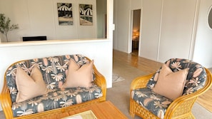 Ein Zimmer mit einer Couch und einem Sessel, beide mit floralem Bezug, einem Korbtisch und einem Spiegel, der den Raum reflektiert.