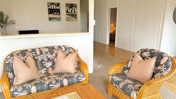 Ein Zimmer mit einer Couch und einem Sessel, beide mit floralem Bezug, einem Korbtisch und einem Spiegel, der den Raum reflektiert.