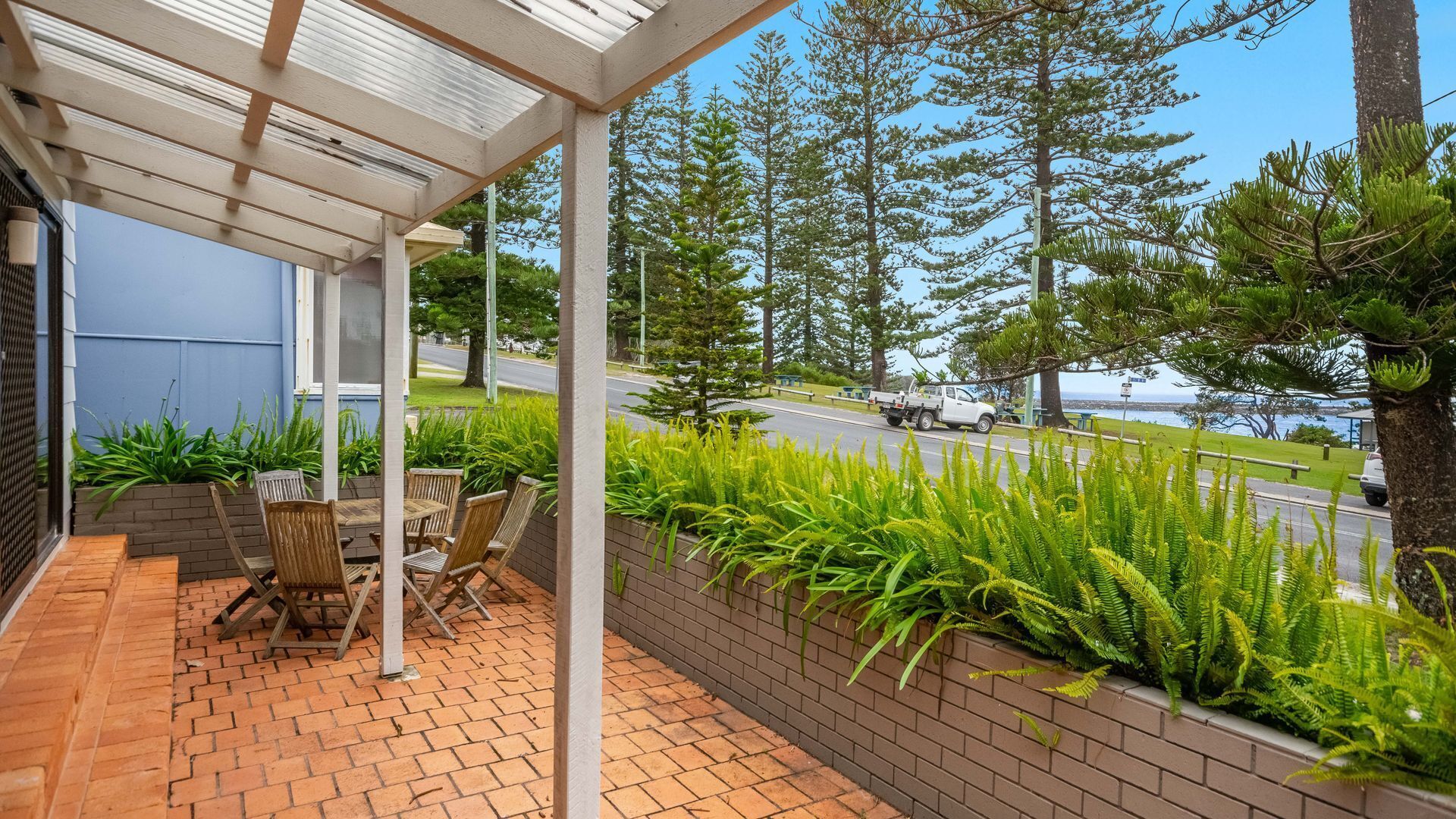 A covered patio with outdoor furniture, a brick planter with ferns, and a view of a road and trees.