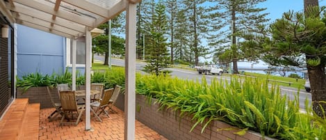 A covered patio with outdoor furniture, a brick planter with ferns, and a view of a road and trees.