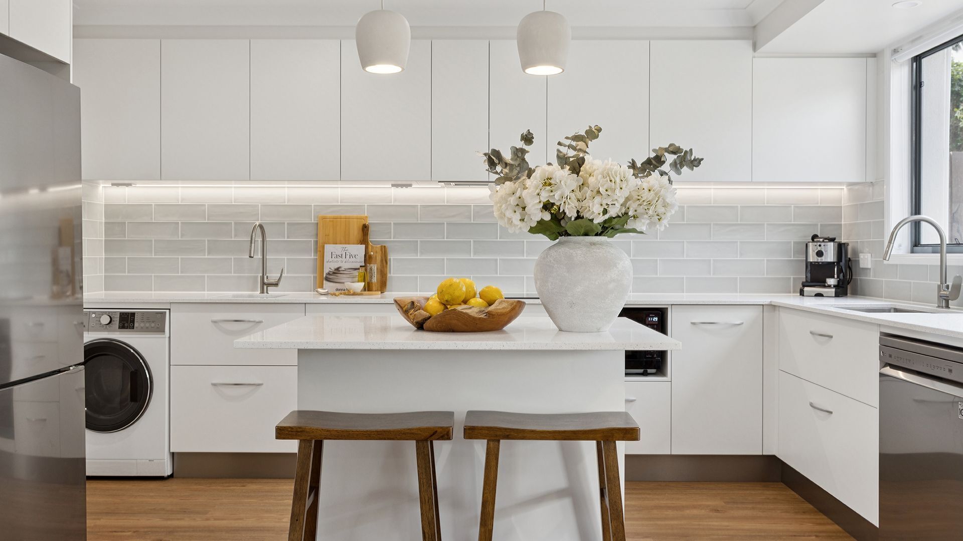 A modern kitchen with white cabinetry, a central island with a vase of flowers, a coffee maker, and a washing machine.
