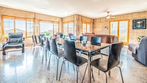 A spacious dining area with a long table and multiple chairs, large windows, and a ceiling fan.