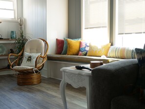 A cozy living room with a wicker chair, a bench with colorful cushions, a small table, and a window with blinds.