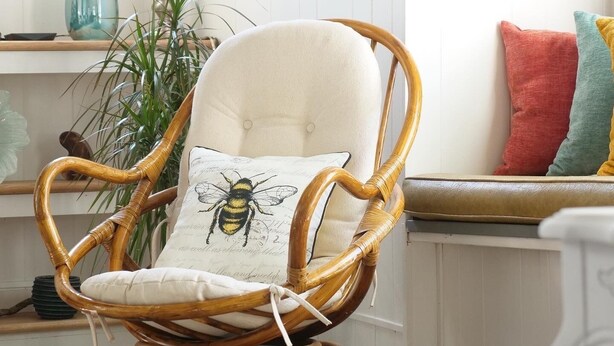 Una silla de mimbre con cojín y almohada con diseño de abeja, ubicada en una habitación con piso de madera y pared blanca.