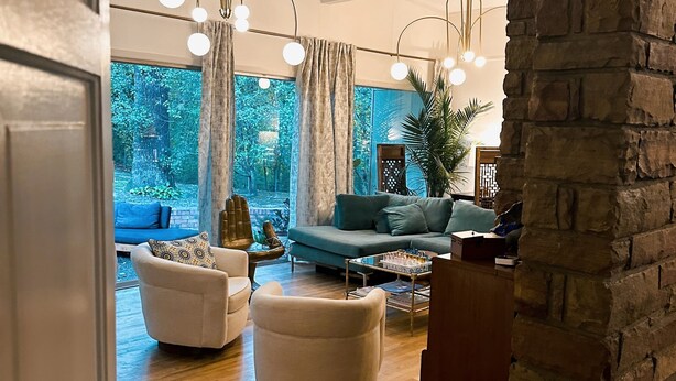 A modern living room with a sofa, armchairs, and a coffee table. There is a large window with curtains, a brick wall, and a hanging light fixture. - Walk to Campus, Minutes to Square: Entire Home w Spacious Yard, Pet-Friendly! (Oxford)