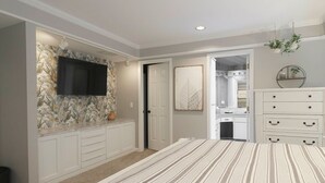 Une chambre avec un grand lit, un téléviseur fixé au mur recouvert d’un papier peint à motifs floraux, une commode blanche et une salle de bain visible à travers une porte ouverte.