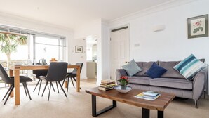 A modern living room with a dining area, a grey sofa with cushions, a coffee table, and a bookshelf. - The Wesley Apartment (Falmouth)