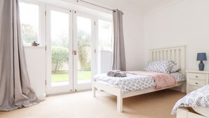 A bedroom with a bed, a nightstand, a lamp, and a view of a garden through a sliding door. - The Wesley Apartment (Falmouth)