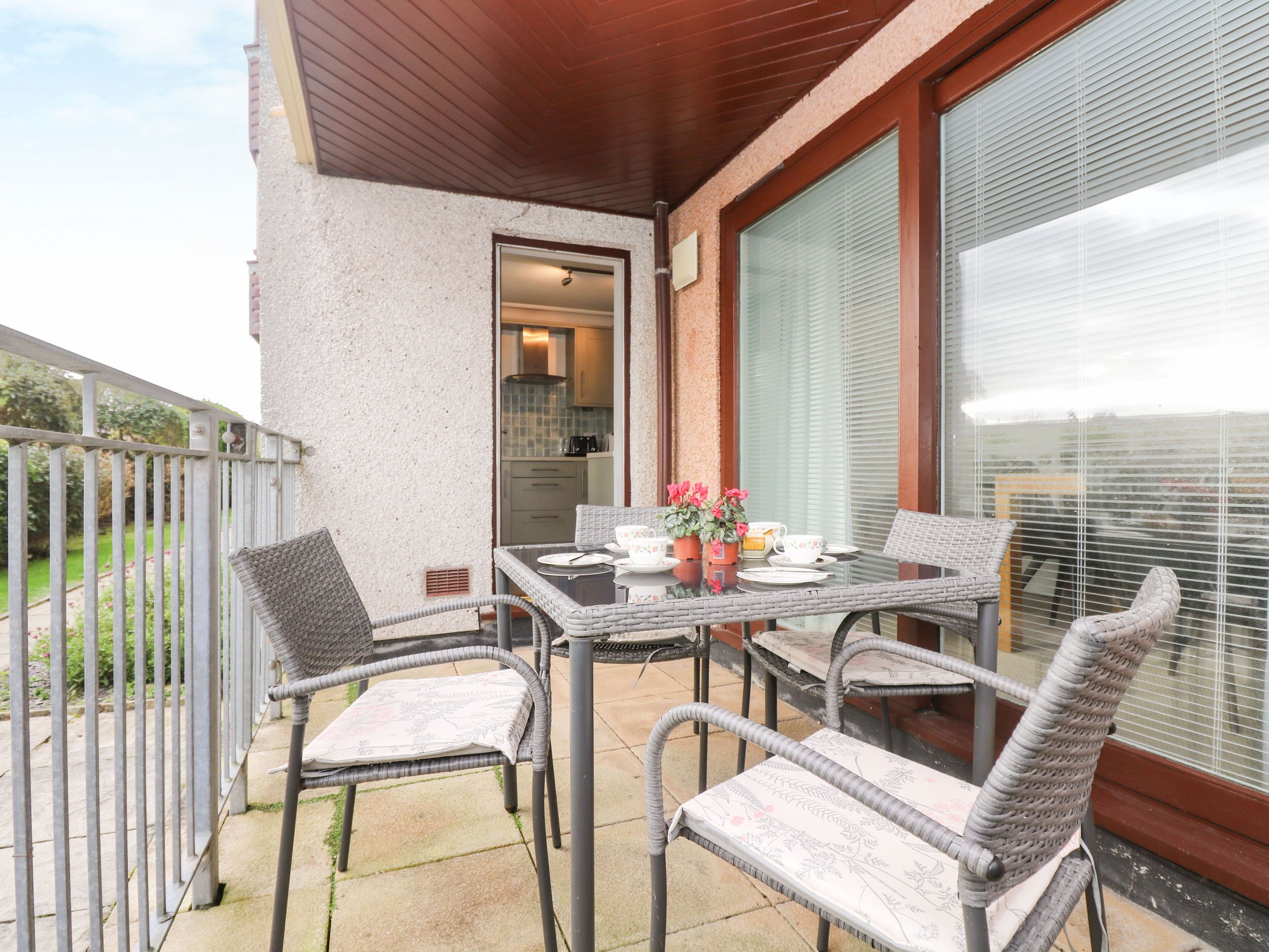 A patio with a table and four wicker chairs, a view of a kitchen, and a large window with blinds.