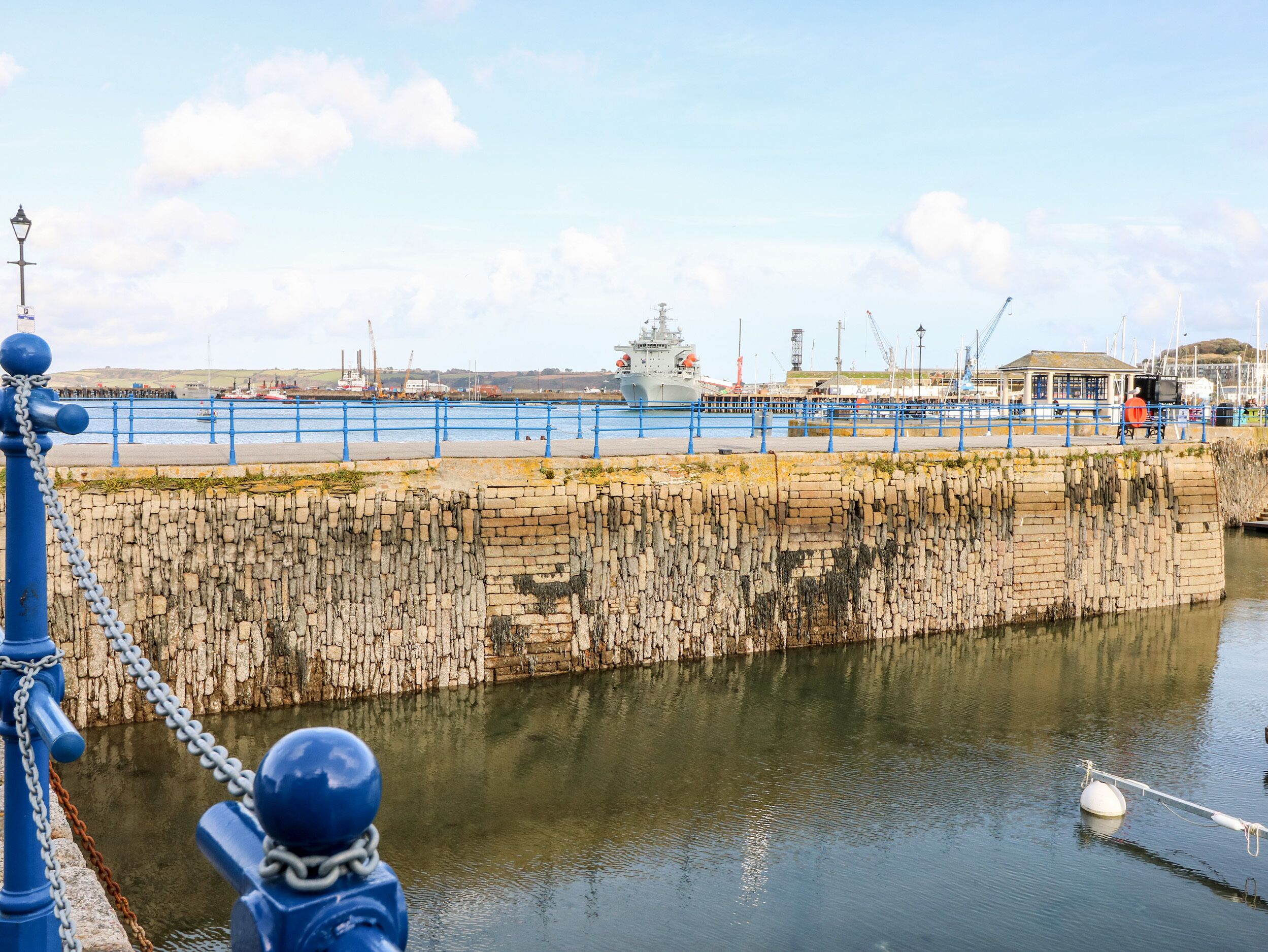 A waterfront with a stone wall, a blue railing, and a ship in the background.