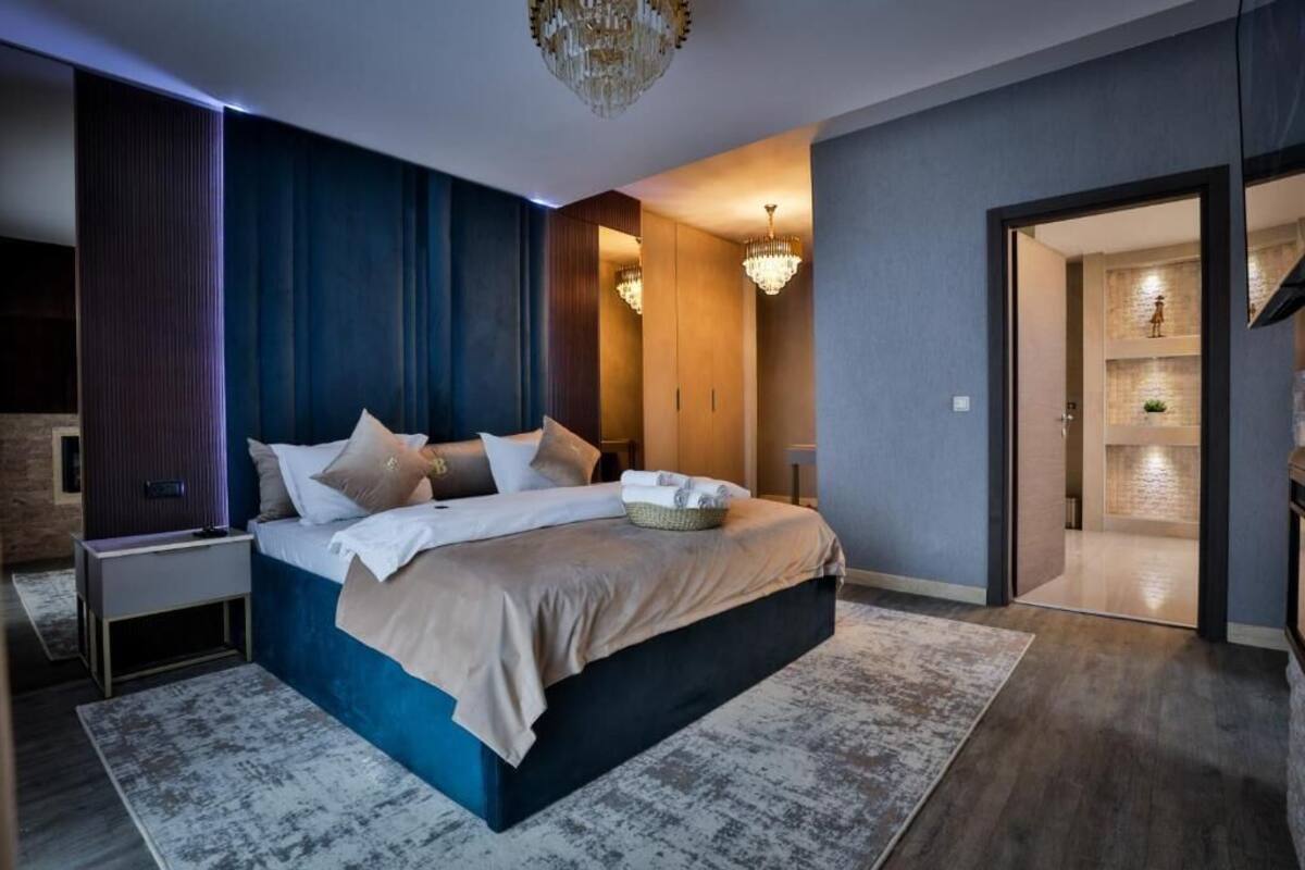 A modern bedroom with a large bed, a nightstand, a chandelier, and a view into another room.