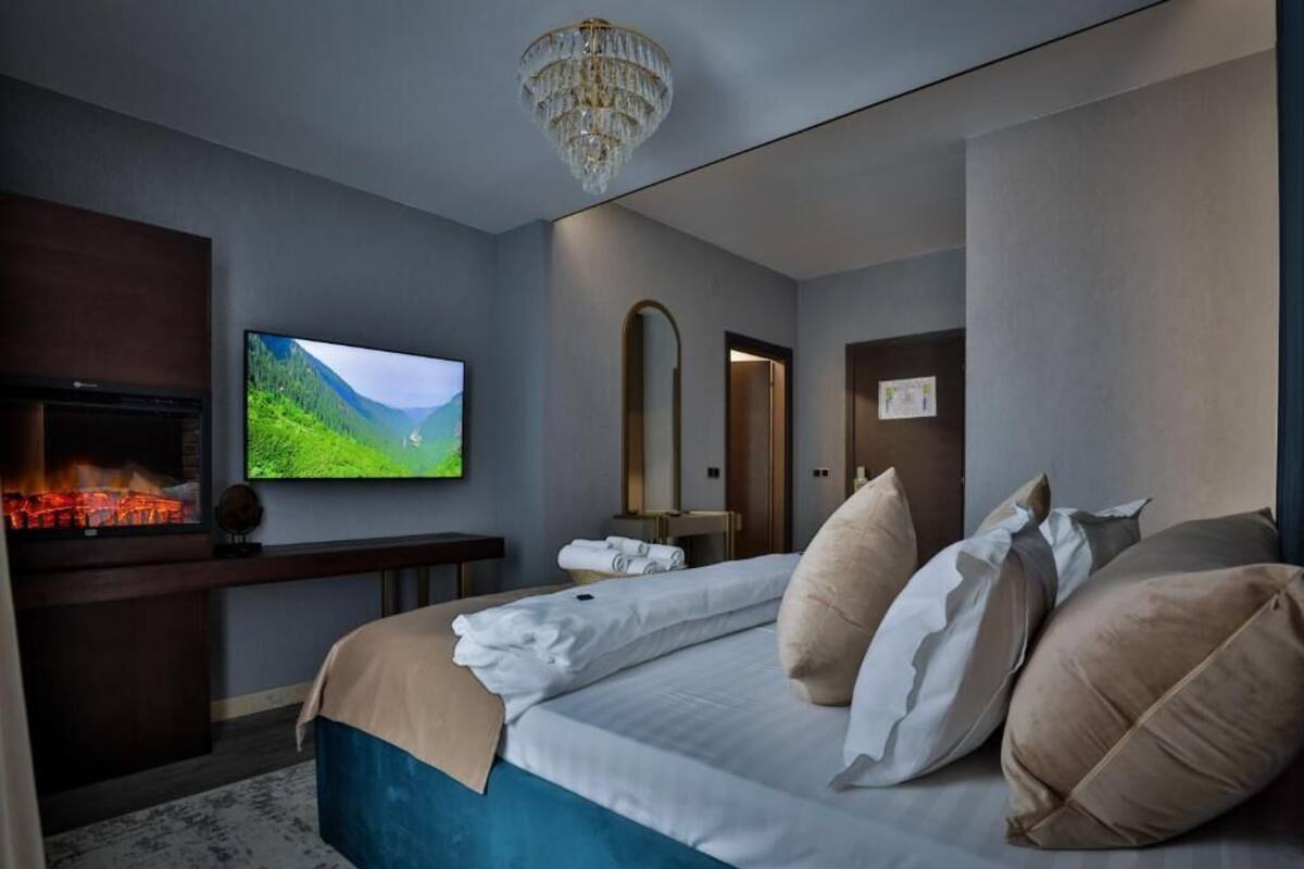 A modern hotel room with a large bed, a flat-screen TV, a fireplace, and a chandelier.