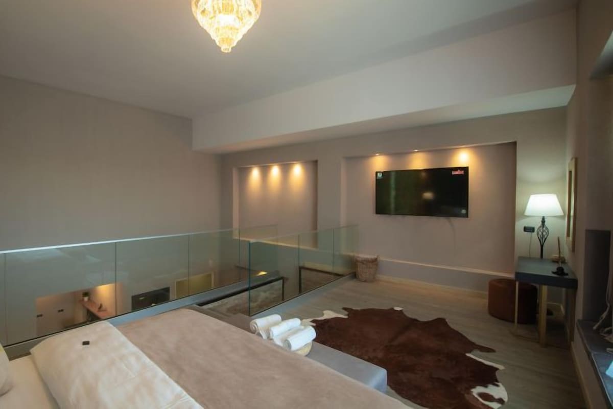 A modern hotel room with a large bed, a glass railing, a flat-screen TV, and a leather ottoman.