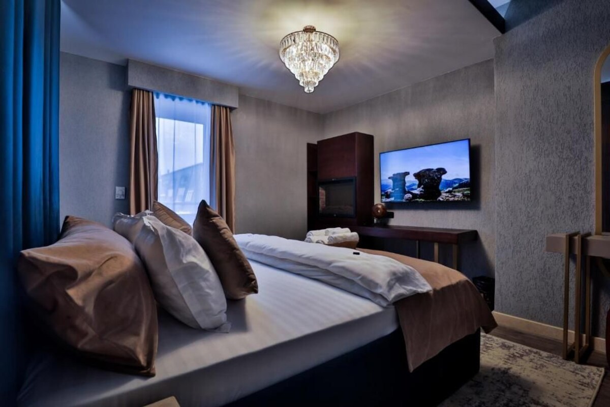A hotel room with a large bed, a television mounted on the wall, a chandelier, and a window with curtains.