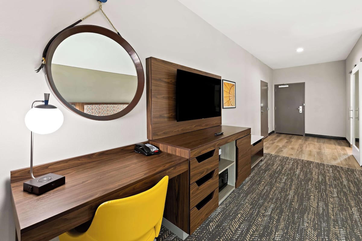 A hotel room with a wooden desk, a round mirror, a flat-screen TV, a yellow chair, a remote control, and a telephone.