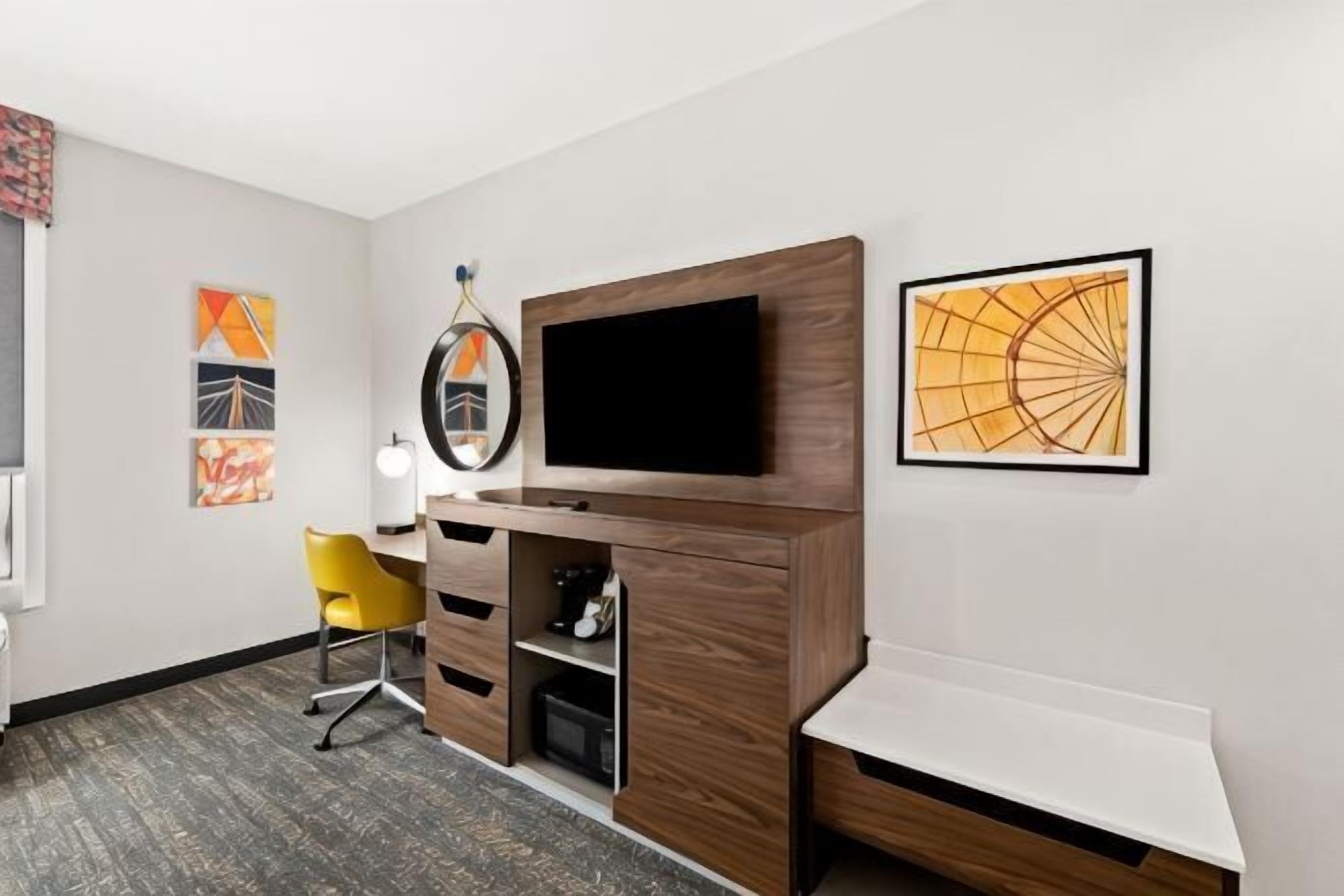 A hotel room with a wooden TV stand, a flat-screen TV, a yellow chair, a desk, and a white bedside table.