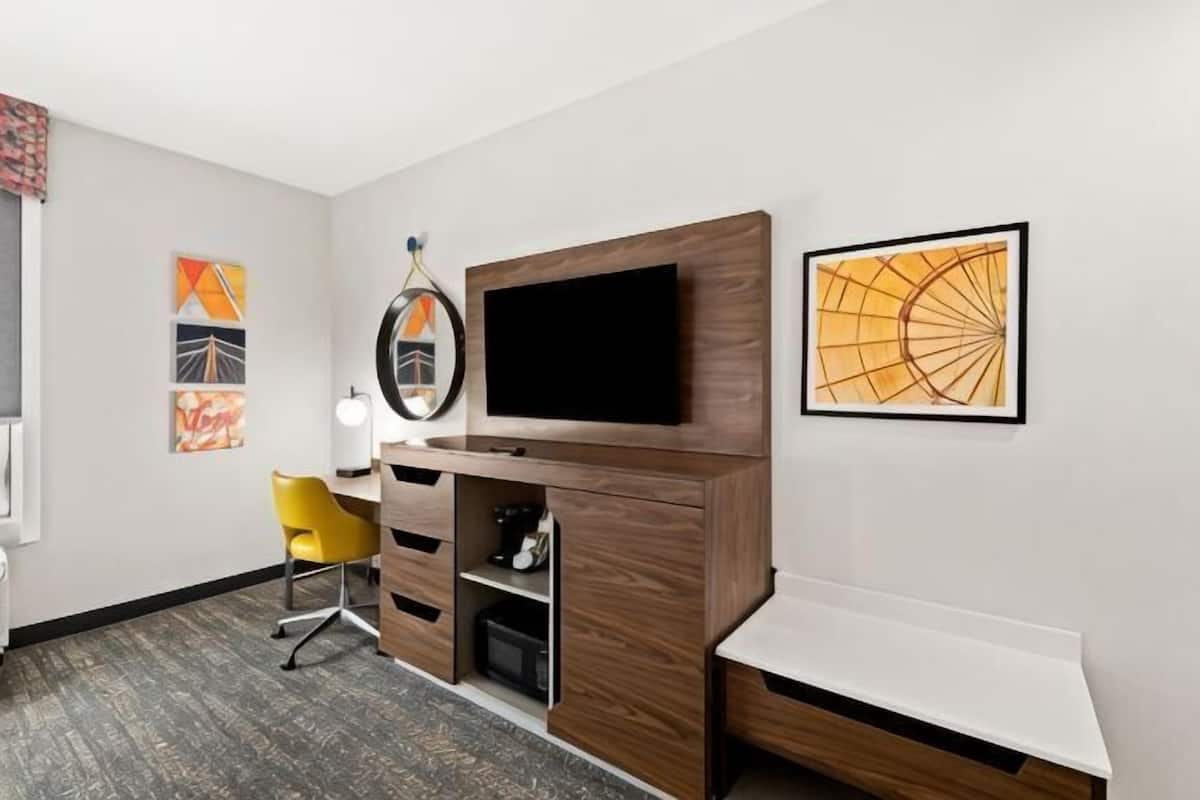 A hotel room with a wooden TV stand, a flat-screen TV, a yellow chair, a desk, and a white bedside table.