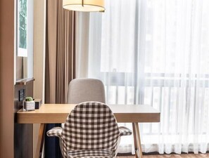 Et hotelværelse med et træskrivebord, en ternet stol, en lampe og lette gardiner.