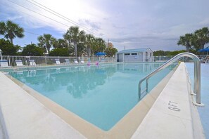 A swimming pool with a metal safety railing, surrounded by a white fence, palm trees, and a small building with a blue roof.