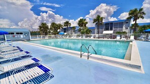 Piscina com espreguiçadeiras, palmeiras e um prédio com telhado azul.