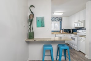 A kitchen with white cabinets, a granite countertop, a microwave, and a stove. There are two blue stools and a green decorative piece on the wall. - B 25 Sundown Condominiums (Pensacola)