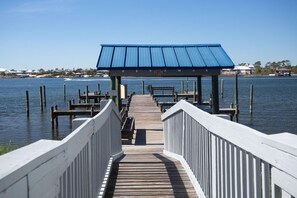 A wooden pier leading to a covered area with a blue roof, surrounded by water and distant buildings.