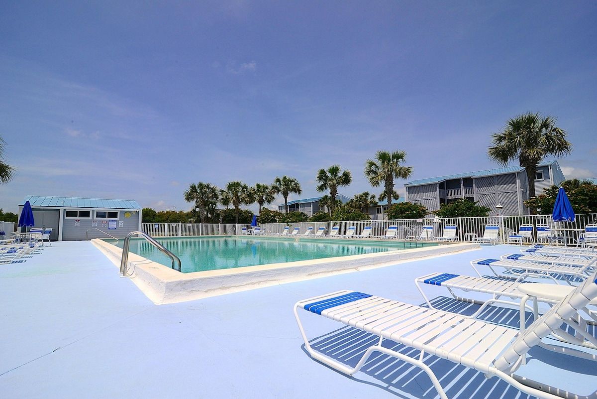 A pool area with lounge chairs and umbrellas, surrounded by palm trees and buildings.
