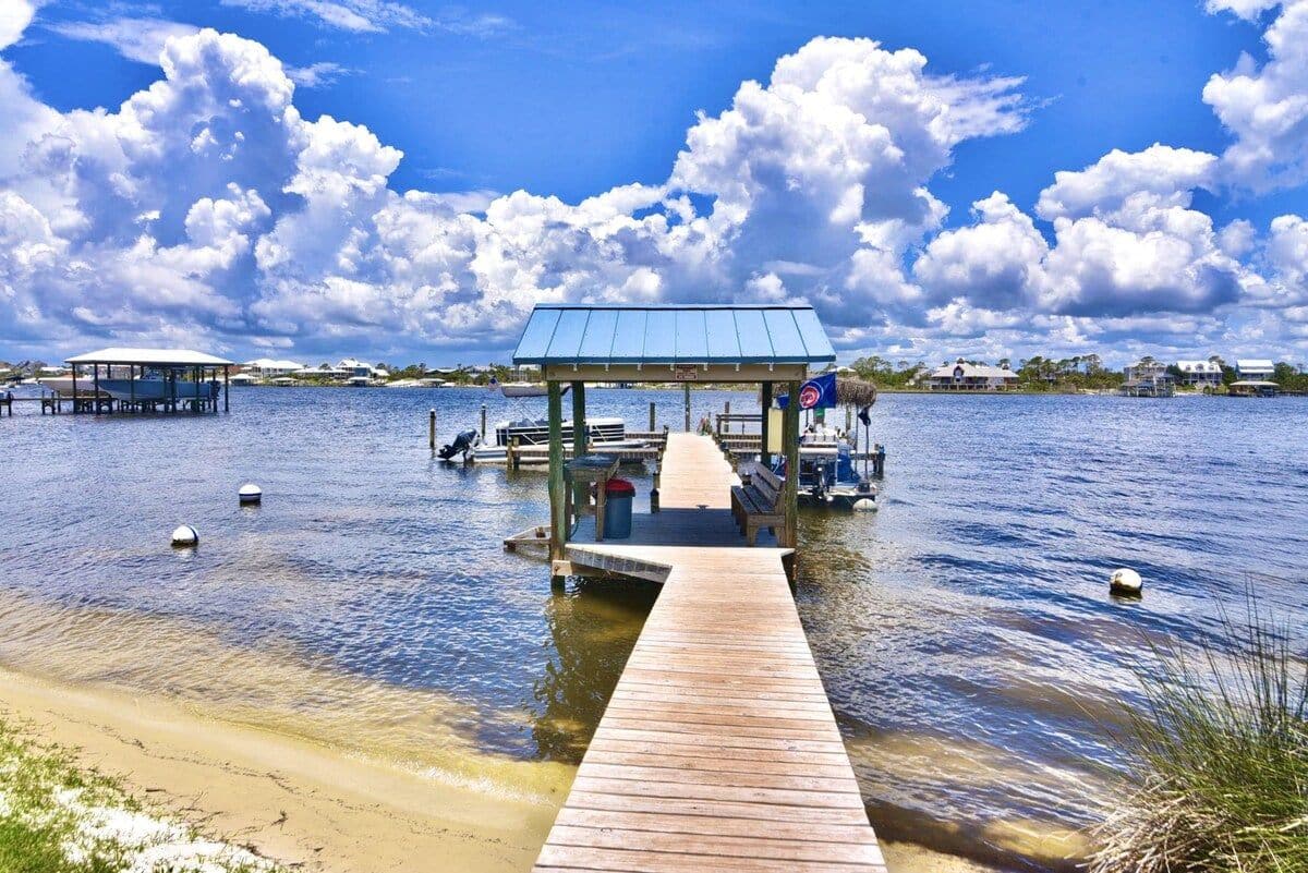 A wooden pier leading to a covered pavilion with benches and a flag, surrounded by calm water and distant buildings.