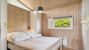 Un dormitorio con techo de madera, una cama con ropa de cama blanca, una ventana con vistas a vegetación y un calentador fijado a la pared.