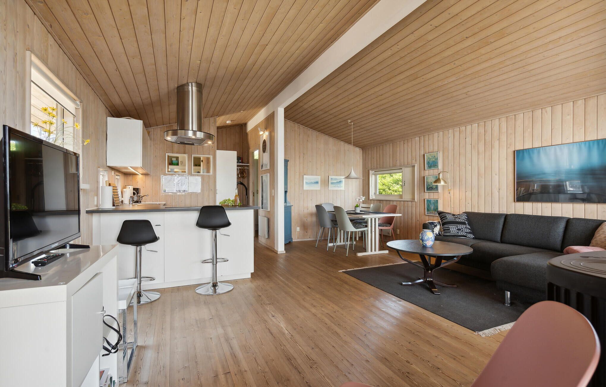 Une cuisine moderne avec un lambris en bois, un îlot central et des tabourets de bar. On y trouve un coin repas avec une table et des chaises, ainsi qu’un salon avec un canapé et une table basse.