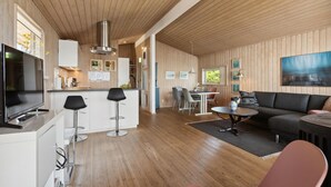 Una cocina moderna con paneles de madera, una isla central y taburetes de bar. Hay una zona de comedor con una mesa y sillas, y una sala de estar con un sofá y una mesita de centro.