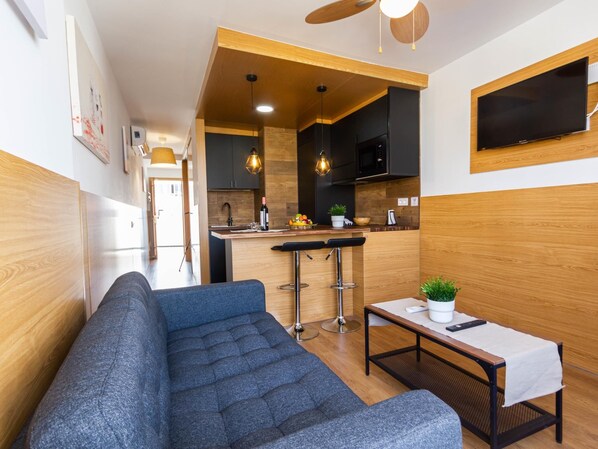 A modern living room with a sofa, a small table with a plant, and a kitchen area with bar stools and a TV.