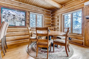 Interior de cabana de madeira com mesa redonda e quatro cadeiras, amplas janelas com vista para a neve e um quadro na parede.