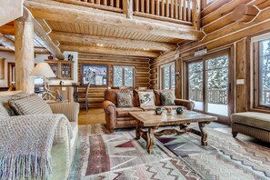 Interior acolhedor de uma cabana de madeira com lareira, móveis de madeira e janelas amplas com vista para o ambiente nevado lá fora.