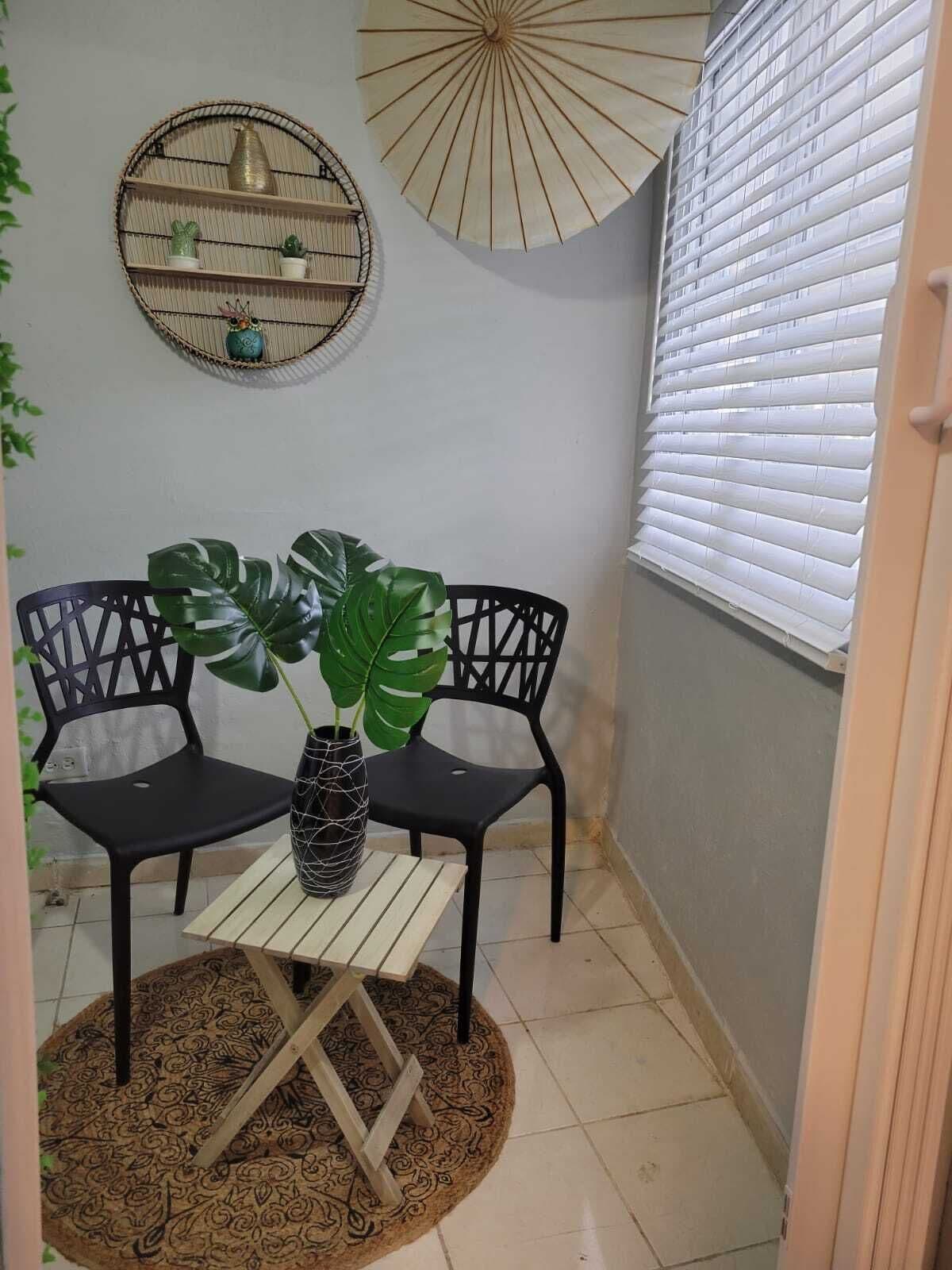 Une pièce d’angle avec deux chaises noires, une petite table sur laquelle est posée une plante, une étagère décorative fixée au mur et un grand ventilateur.