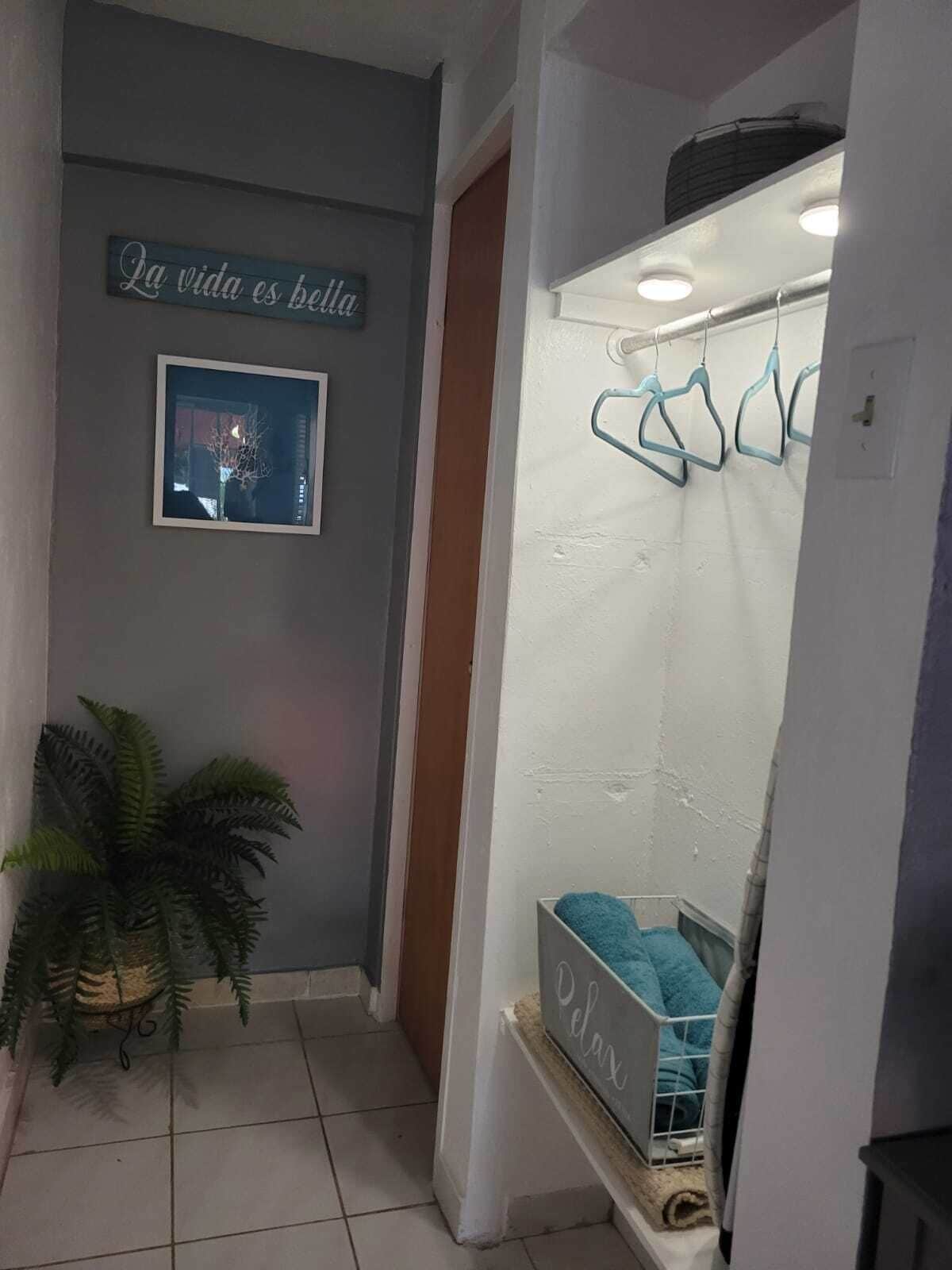 A hallway with a sign reading "La vida es bella," a framed picture, a potted plant, and a relaxation area with a towel and a basket.