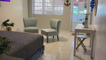 A bedroom with a bed, two chairs, a small table, a TV, and a window with blinds.