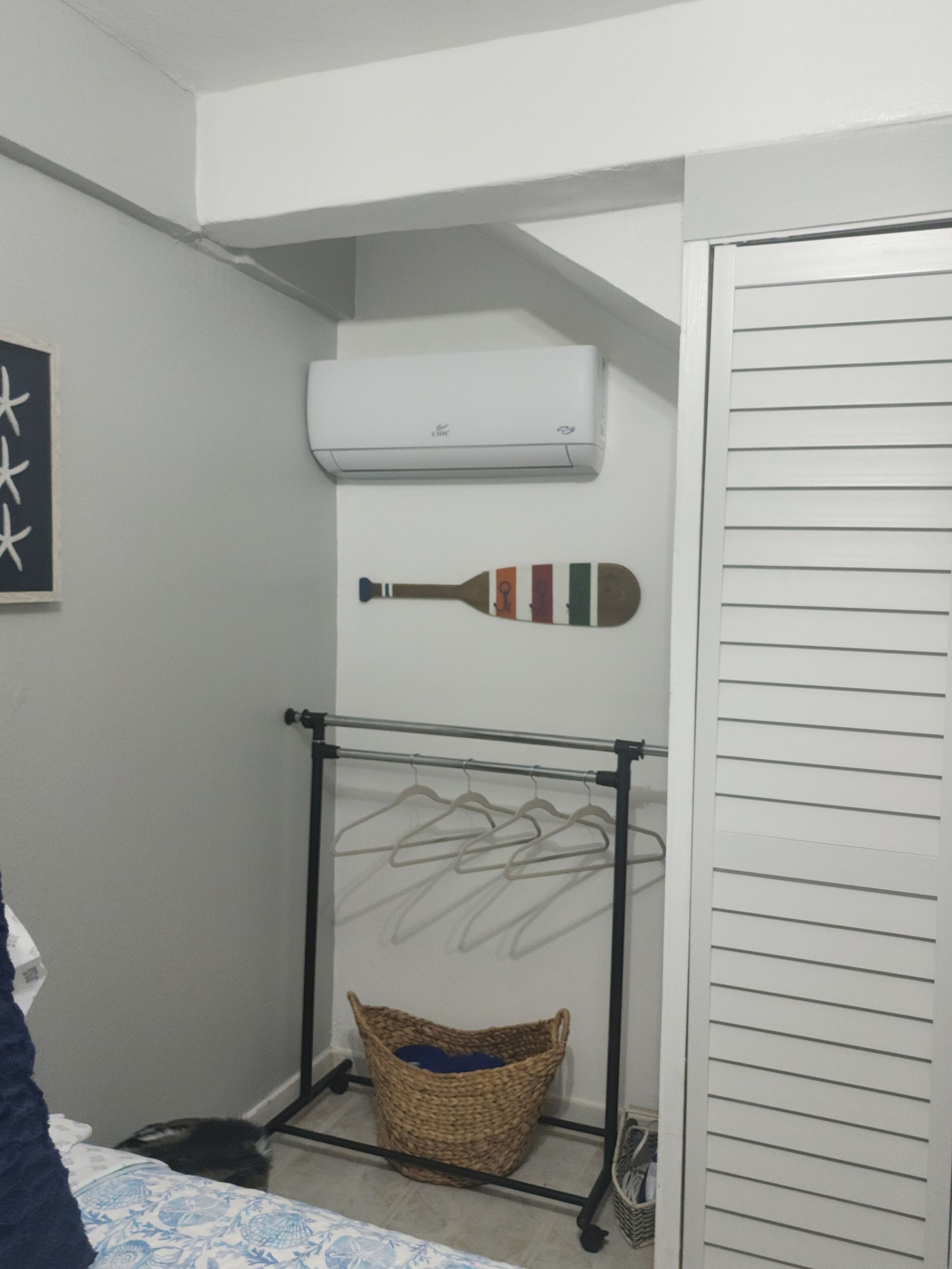 A small room with a bed, a wall-mounted air conditioner, a drying rack with clothes, and a basket.