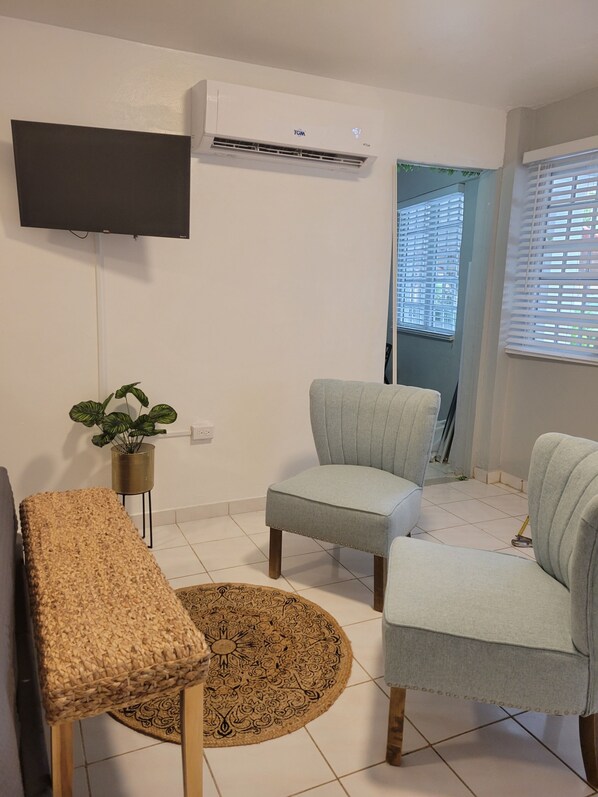 Una sala de estar con un televisor de pantalla plana montado en la pared, un sillón gris, un banco tejido, un tapete estampado y una planta en maceta.