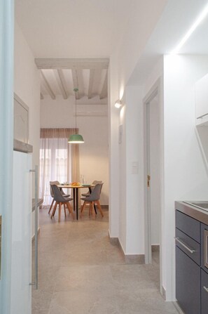 Un couloir étroit mène à un espace repas comprenant une table et des chaises, un luminaire suspendu et un coin cuisine avec des placards.