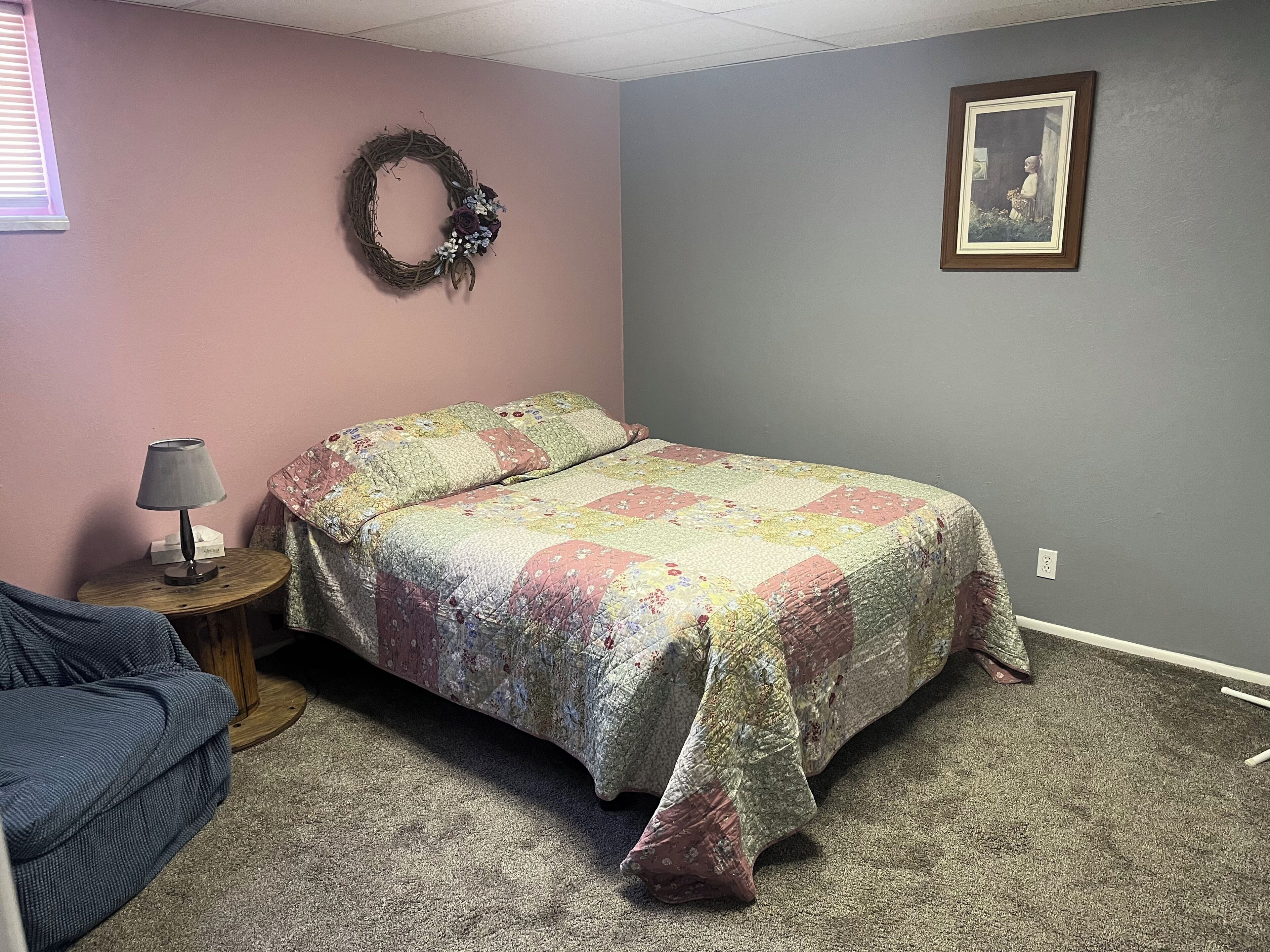 A bedroom with a bed, a nightstand, a lamp, a chair, a wall decoration, and a framed picture.