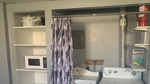 A compact laundry room with a washing machine, dryer, and microwave. There are shelves above the appliances, and a curtain partially covers the doorway. - Grandma Marva’s Hideout basecamp for many National & State Parks (Blanding)