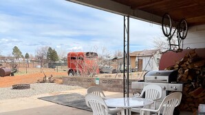 A covered patio area with a table and chairs, a grill, and a view of a red truck and buildings in the background. - Grandma Marva’s Hideout basecamp for many National & State Parks (Blanding)