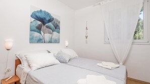 Ein Schlafzimmer mit Bett, weißer Bettwäsche, einem Wandbild, einem Nachttisch mit Lampe und einem Fenster mit durchsichtigen Vorhängen.