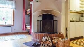 A living room with a fireplace, a window with curtains, a kitchen area, and a wooden floor.