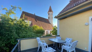A wooden deck with a table and chairs, a yellow building, and a church with a tower in the background. - Vacation domicile - Comfortable vacation apartment OG1 for 1-5 pers. Central. Terrace. (Bad Radkersburg)