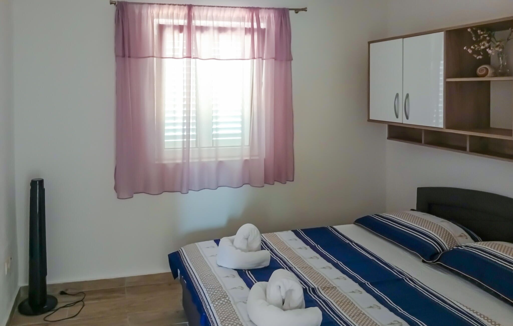 A bedroom with a bed, a window with pink curtains, a standing fan, and a cabinet with a mirror.